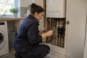 A woman heating engineer crouches beside an open boiler cupboard in a UK home, holding a wrench and checking her phone